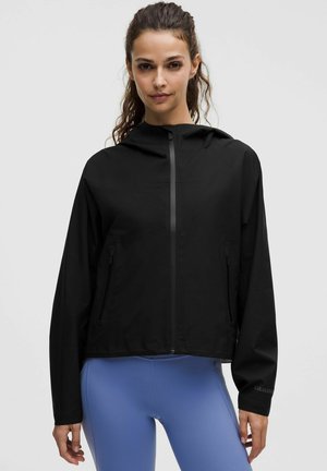 Frau mit langen Haaren, die eine schwarze Kapuzenjacke mit Reißverschluss und blaue Leggings trägt, steht vor einem schlichten, hellen Hintergrund.