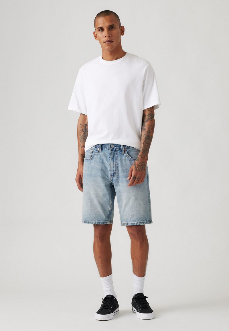 Tricou alb din bumbac, pantaloni scurți din denim albastru deschis și adidași negri cu accente albe. Stilizat cu șosete scurte, evidențiind un aspect casual.