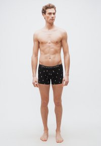 Νέος άνδρας στέκεται ξυπόλητος φορώντας μαύρα boxer Ralph Lauren Polo με λευκά λογότυπα, μπροστά σε απλό φωτεινό φόντο.