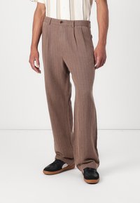 Pantalones marrones a rayas con un corte relajado, pliegues en la parte delantera y una textura suave. Combinados con zapatillas negras con suela de goma.