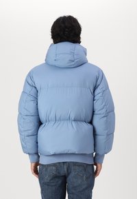 Giacca puffer azzurra chiara con colletto alto, imbottitura spessa e polsini a coste. Presenta un cappuccio e una vita aderente. Design minimalista.