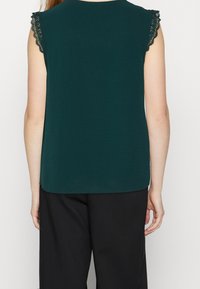 Dark green sleeveless top with lace trim on shoulders, smooth texture, rounded hem, paired with black trousers.