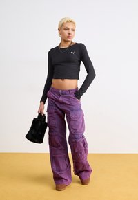 Top cropped preto de manga longa com um logótipo branco; calças cargo roxas com vários bolsos; modelo a segurar uma pequena mala de pelo sintético preto.