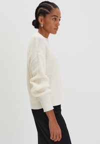 Maglione a maglia crema con motivo testurizzato, spalle calate e polsini a costine. Abbinato a pantaloni neri.