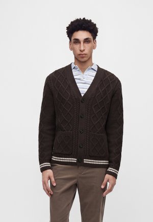 Mann trägt eine dunkelbraune Strickjacke mit Zopfmuster über einem weißen, gestreiften Poloshirt und hellbraune Hose, steht vor einem einfarbigen Hintergrund.