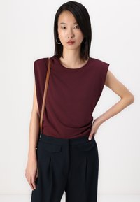 Burgundy short-sleeve top with structured shoulders, paired with dark pleated trousers. The model holds a brown shoulder bag.