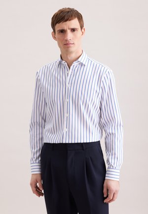 Homme portant une chemise blanche à manches longues avec des rayures verticales bleues et un pantalon sombre plissé, debout devant un fond uni.