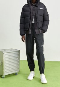 Personne portant une doudoune noire, une chemise sombre, un jogging Nike noir et des baskets blanches, debout à côté d'une boîte de rangement en métal argenté sur roulettes.