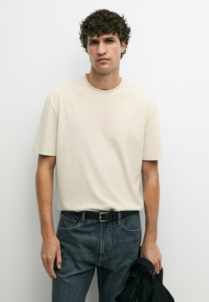 Jonge man met krullend donker haar, een eenvoudige beige t-shirt die in een donkerblauwe spijkerbroek is gestopt, en een zwart jack in zijn hand houdend.