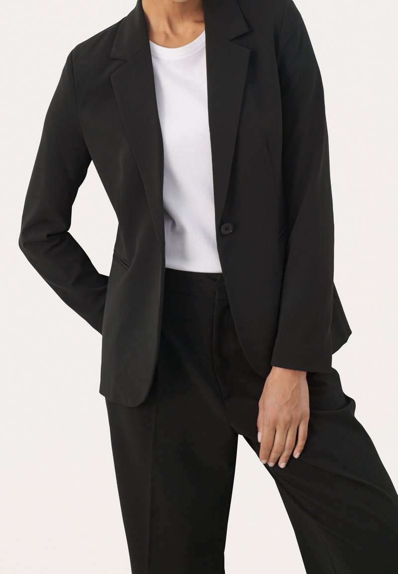 Personne portant un blazer noir à un bouton unique et un pantalon noir assorti par-dessus un t-shirt blanc uni, sur un fond blanc.