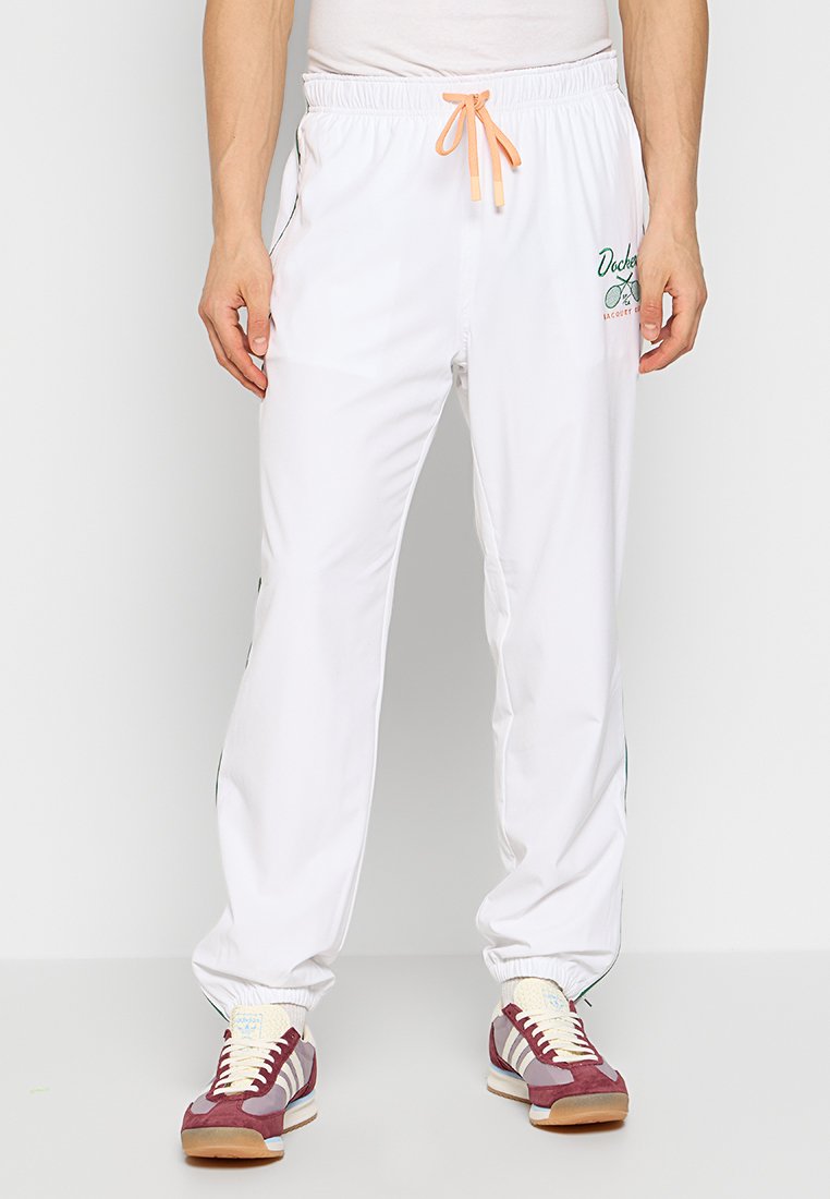 Witte sportbroeken met een elastische tailleband en oranje trekkoord. Bevat een groen logo detail aan de linkerkant en zwarte zijstrepen.