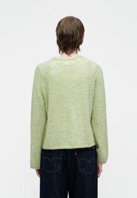 Person med kort brunt hår, iført en lys grønn langermet genser og mørkeblå jeans, som står mot en enkel hvit bakgrunn.