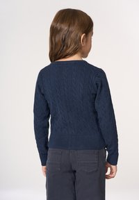 Maglione blu navy a lavorazione a trecce con scollo rotondo e polsini e orlo a coste, indossato con pantaloni grigi. Il retro presenta un motivo testurizzato.