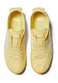 Żółte płócienne trampki z białymi akcentami, zaokrąglony czubek, minimalistyczny design, brak sznurowadeł z przodu, teksturowana tkanina i metka z logo na języku.