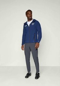 Chaqueta navy con cremallera y capucha, con un logo, combinada con pantalones grises ajustados. Ambos artículos tienen un diseño elegante y un ajuste atlético. Calzado negro.