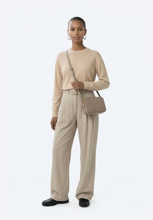Nainen, joka pukeutunut beigeen neulepuseroon ja korkeavyötäröisiin laskostettuihin housuihin, mustiin loafer-kenkiin, renkaanmuotoisiin korvakoruihin ja kantaa harmahtavanruskeaa olkalaukkua.