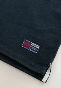 Navyblaues Gewebe mit einem Superdry-Label, das "Vintage Athletic Outfitters" liest, mit detaillierten Nähten und einem kleinen seitlichen Schlitz.