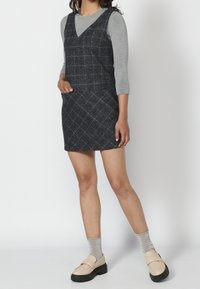 Navy plaid pinafore dress with a V-neck, sleeveless design, front pocket, paired with a gray long-sleeve shirt and beige loafers.