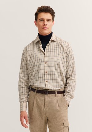 Hellbeige karierte Hemd mit Holzknöpfen, kombiniert mit beigen strukturierten Hosen. Über einem dunklen Rollkragenpullover getragen, um Lagen zu schaffen.