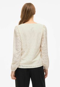 Kremowa bluzka z fakturowanym wzorem w diamenty, długimi bufkami i prostym dekoltem. Materiał wydaje się lekki i przewiewny.