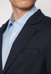 Navy blazer made of textured fabric, featuring notched lapels, a flap pocket, and worn over a light blue striped dress shirt.