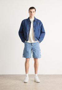 Giacca in denim con colletto e zip frontale, abbinata a pantaloni corti in denim blu chiaro. Il modello indossa sneakers bianche e calze. Look semplice e casual.