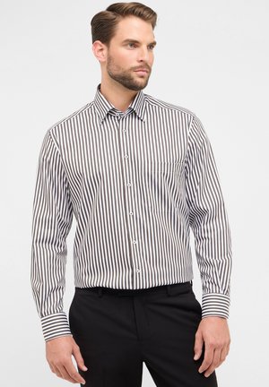 Uomo che indossa una camicia bianca a maniche lunghe con righe verticali marroni e pantaloni neri, in piedi contro uno sfondo chiaro e uniforme.