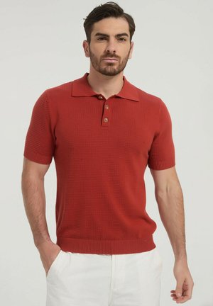 Homme aux cheveux courts et foncés avec une barbe, portant un polo en maille à manches courtes couleur rouille et un pantalon blanc, debout avec une main dans la poche.