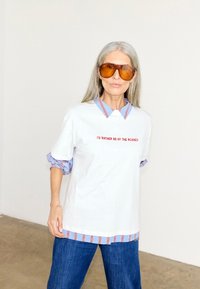 Ηλικιωμένη γυναίκα με μακριά γκρίζα μαλλιά, φοράει μεγάλα πορτοκαλί γυαλιά ηλίου, λευκό πουκάμισο πάνω από ριγέ πουκάμισο με γιακά και μπλε τζιν.