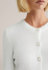 Weißer gestrickter Cardigan mit weichem Material, rundem Kragen und drei hexagonalen Knöpfen mit goldfarbener Umrandung.