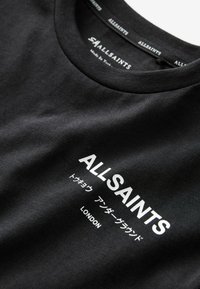 Juodas medvilnės marškinėliai su apvaliu iškirpimu, ant kurių balta spalva užrašyta "ALLSAINTS" ir "LONDON" stora šriftu, o žemiau pateikti japonų simboliai.
