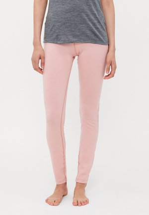 Person som har på seg lyserosa, tettsittende tights og en grå, avslappet topp, som står barføtt foran en ensfarget hvit bakgrunn.