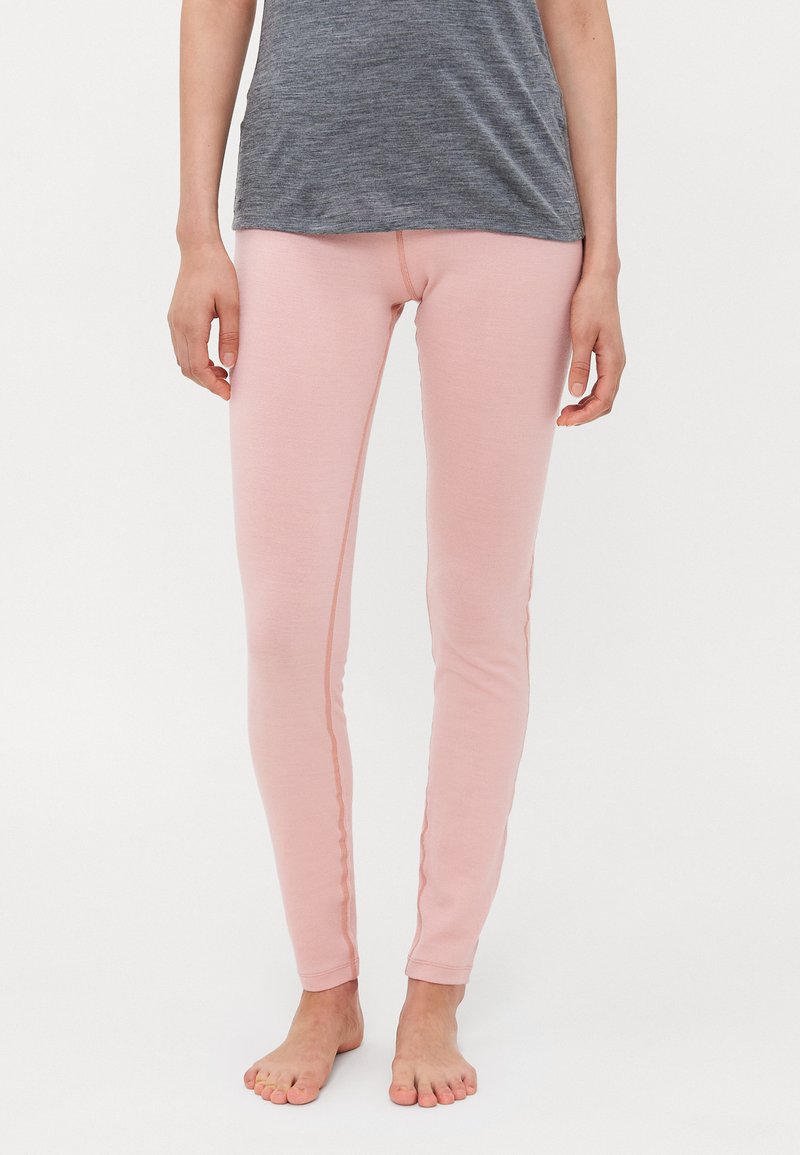 Person som har på seg lyserosa, tettsittende tights og en grå, avslappet topp, som står barføtt foran en ensfarget hvit bakgrunn.