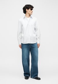 Férfi fekete napszemüvegben, fehér gombos ingben, bő kék farmernadrágban és fekete cipőben, sima világos háttér előtt állva.