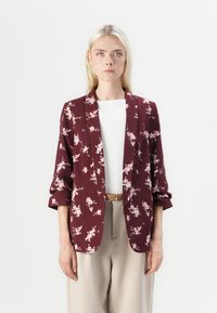 Blazer floreale borgogna con maniche arrotolate, realizzato in tessuto leggero. Presenta un colletto a risvolto e una vestibilità comoda, indossato sopra una maglietta bianca.