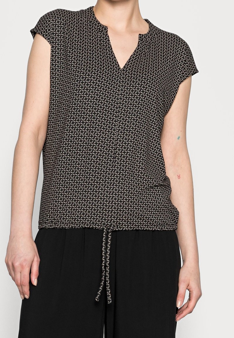Svart och vit mönstrad kortärmad topp med V-ringning och knytning i midjan, tillverkad av lätt tyg. Texturerad geometrisk design.