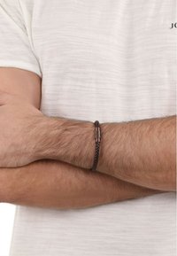 Bracciale in pelle intrecciata marrone con chiusura in metallo, indossato su un polso contro una camicia a trama chiara. Notabile il motivo di tessitura prominente.