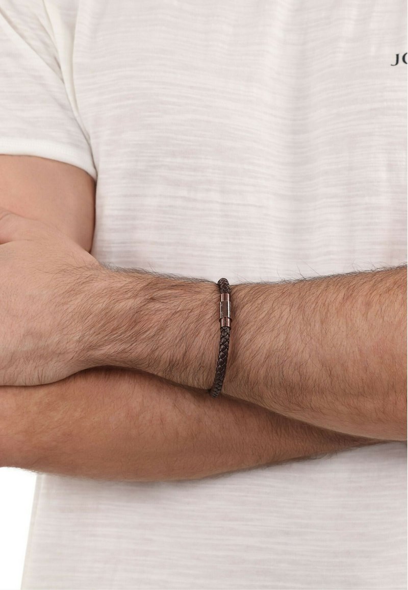 Bracciale in pelle intrecciata marrone con chiusura in metallo, indossato su un polso contro una camicia a trama chiara. Notabile il motivo di tessitura prominente.