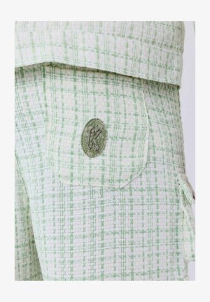 Groen en witte textuurstof, met een patch zak voorzien van een grijze K-embleem. De stof heeft een geweven patroon, wat visuele diepte creëert.