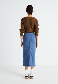 Rjav rebrast pulover z zavihanimi rokavi, v kombinaciji z midi jeansom v svetlo modri barvi. Model nosi bele nogavice in črne čevlje.