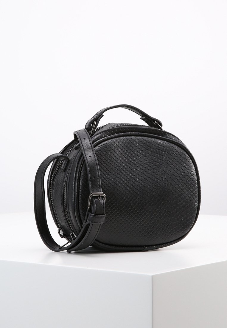Sac à bandoulière circulaire noir en cuir texturé, avec fermeture éclair, une sangle amovible et une poignée supérieure pour le port.