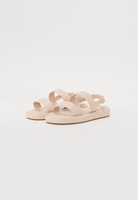 Beige sandals with two wide, crossed straps and a thin ankle strap, featuring a slight heel and smooth, matte finish.