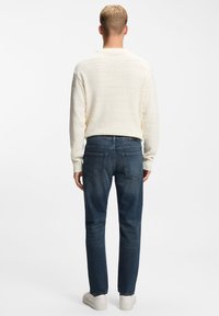 Suéter texturizado de color beige claro combinado con vaqueros de corte recto azul oscuro y zapatillas blancas. La vista es desde atrás, destacando el ajuste.