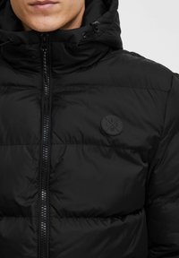 Black padded jacket featuring a zip closure and drawstring hood. Includes a round, black logo patch on the upper left side.