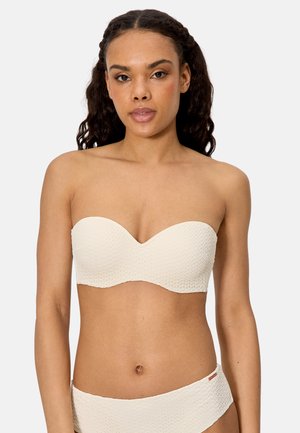 Mujer con cabello oscuro y ondulado que lleva una blusa bandeau crema texturizada sin tirantes y pantalones altos a juego, posando sobre un fondo liso.