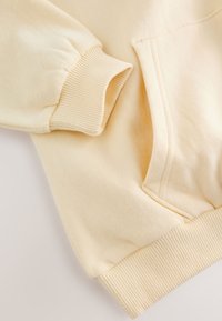 Samtfarbenes Sweatshirt mit geripptem Bündchen an den Ärmeln und passendem geripptem Saum neben einer Kängurutasche aus weichem Stoff.