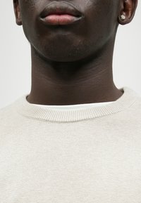 Maglione a maglia beige chiaro con colletto a coste, indossato sopra una maglia bianca. Primo piano delle caratteristiche del collo e della parte inferiore del viso.