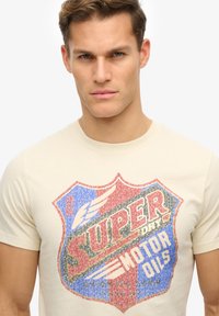 Camiseta beige con un gráfico de escudo colorido que incluye elementos rojos, azules y blancos, y el texto "SUPER DRY MOTOR OILS" en un estilo vintage.