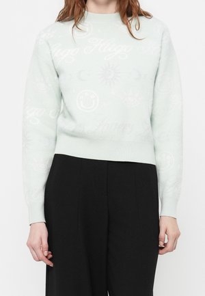 Frau trägt einen hellblauen Pullover mit dezenten Mustern von Smiley, Sonne, Mond und "Hugo" kombiniert mit hoch taillierten schwarzen Hosen.