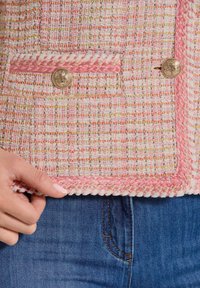 Rosa und weiße Tweedjacke aus strukturiertem Stoff, mit goldenen Knöpfen, geflochtenem Besatz und zwei vorderen Taschen. Kombiniert mit blauer Jeans.